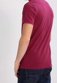Burgundy bomullst-shirt med kort ärm, figurnära design, slät textur och rundad halsringning, bärs tillsammans med mörkblå jeans.