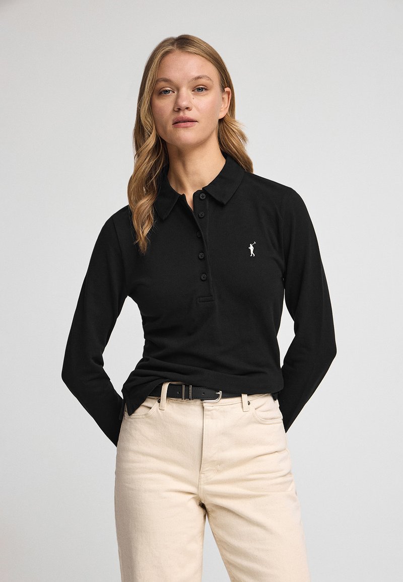 Camisa polo negra de manga larga con cuello, con una tapeta de botones y un pequeño logotipo blanco. Combinada con pantalones beige claros.