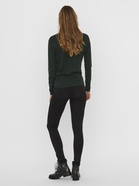 Donna con lunghi capelli rossi che indossa un maglione verde scuro con texture, jeans skinny neri e stivali militari neri, in piedi di spalle.