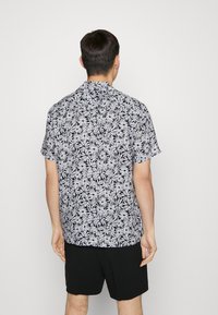 Hombre de espaldas con una camisa de manga corta de patrón floral blanco sobre fondo negro y pantalones cortos negros lisos, frente a un fondo neutro.