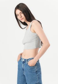 Halli lõhiku ja ümmarguse kaelusega hall crop top riberitud kangas, koos kõrge vöökohtadega siniste teksadega, käed taskutes.