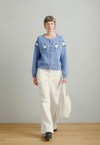 Ανοιχτό μπλε πλεκτή ζακέτα με κεντήματα λουλουδιών, μεγάλα κουμπιά· συνδυασμένη με λευκό παντελόνι και κρεμ τσάντα tote· μαύρα παπούτσια.