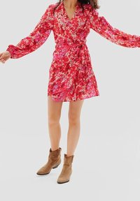Femme portant une robe portefeuille rouge à fleurs avec manches longues et des bottines beige, debout les bras tendus devant un fond gris.