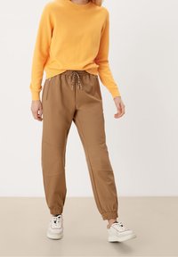 Brune joggerbukser med elastiske manchetter, snøre i taljen og sidelommer med lynlås. Sat sammen med en gul sweater. Sneakers fuldender outfittet.