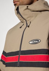 Μπεζ ζακέτα με κο�υκούλα, με μπροστινό μισό φερμουάρ, διακοσμημένη με μαύρες και κόκκινες οριζόντιες ρίγες και κεντημένο λογότυπο στο στήθος.