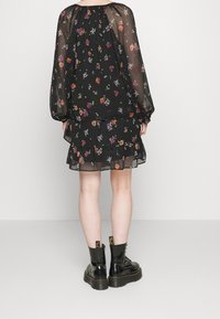 Mujer vestida con un vestido largo de flores negras y mangas transparentes, con botas de plataforma negras y cordones, de pie de espaldas sobre un fondo liso.