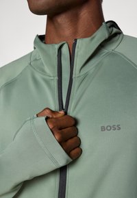 Green athletic zip-up jacket with a high collar, featuring black zip detail and branding on the chest. Fabric has a smooth texture.