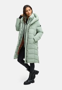 Lichtgroene pufferjas met een lange uitstraling, gequilt oppervlak, oversized capuchon en zwarte accenten. Gecombineerd met een grijze hoodie en zwarte jeans.