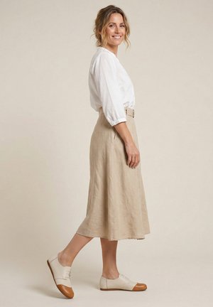 Vrouw glimlacht, loopt zijwaarts en draagt een witte blouse, een beige midi-rok en beige en bruine oxford schoenen tegen een effen achtergrond.