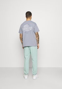 Man med tatuerade armar bär en ljuslila t-shirt med texten "No Thanks", mintgröna joggers och vita sneakers, stående vänd mot en vit vägg.