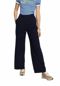 Vidbenna marinblå byxor i bomullsblandning, med hög midja, framfickor och en mjuk textur. Kombineras med beiga sneakers.