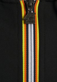 Veste noire à fermeture éclair centrale avec des rayures colorées : jaune fluo, bleu marine, orange et blanc. Détails en métal.