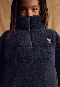 Pull en polaire bleu marine avec un col haut, une demi-zip à l'avant et un petit logo brodé sur la poitrine. Tissu doux et texturé.