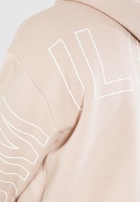Haut à capuche rose clair avec un design de texte blanc surdimensionné dans le dos et des motifs de lignes abstraites. Tissu doux avec une texture lisse et une coupe décontractée.