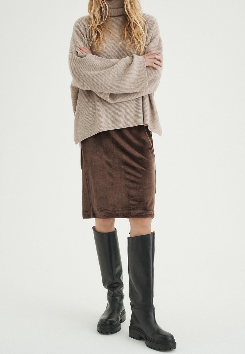 Femme aux longs cheveux blonds portant un pull à col roulé beige oversize, une jupe marron jusqu'aux genoux et des bottes noires montantes, debout les bras croisés.