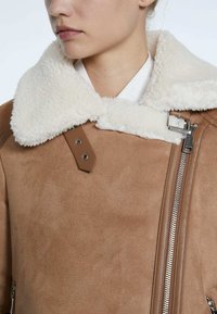 Close-up van een persoon in een tan schapenvachtjas met een grote witte fleece kraag en zilveren rits- en gespdetails.