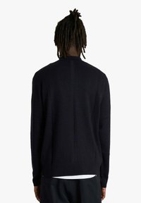 Persona con dreadlocks che indossa un maglione nero a maniche lunghe, vista da dietro su uno sfondo bianco uniforme.