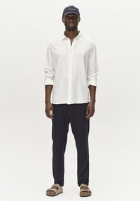 Weißes Leinenhemd mit Knöpfen, marinefarbene Hose, beigefarbene Sandalen und eine marineblaue Kappe mit Text. Das Outfit zeichnet sich durch eine lockere Passform, leichte Stoffe und neutrale Farben aus.