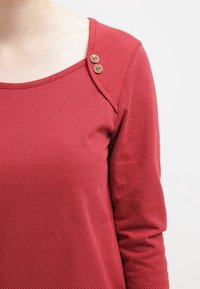 Röd långärmad topp med rundad halsringning, textiler med små prickar och två dekorativa knappar på vänster axel.