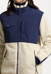 Fleece jack met een marineblauwe bovenkant en een crèmekleurige onderkant. Voorzien van ritssluiting, zijzakken en een hoge kraag. Zachte textuur.