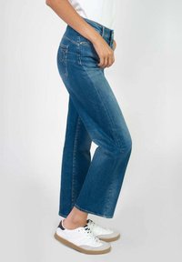 Vaqueros de mezclilla azul de cintura alta con corte recto, que presentan dos bolsillos traseros y detalles de costura dorada. Combinados con zapatillas blancas.