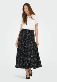 Schwarzer maxikleid im floralen Muster mit weißen Motiven, aus leichtem Stoff. Kombiniert mit einem weißen T-Shirt und schwarzen Ballerinas mit Schleifen.