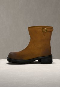 Bottine en cuir marron avec semelle noire et petit talon, avec une bride latérale et un détail de boucle dorée.