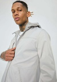 Veste zippée gris clair avec capuche, présentant une texture lisse, des poignets élastiques et une poche avant. Design simple, sans motifs visibles.