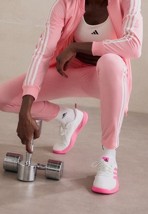 Osoba ubrana w różowy sportowy dres i biało-różowe sneakersy, kuca, trzymając srebrną hantlę na jasnej podłodze.
