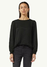 Schwarze Langarmbluse mit Hahnentrittmuster, Rundhalsausschnitt, strukturierter Stoff und gerafften Bund, kombiniert mit dunklen Jeans.