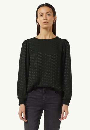 Schwarze Langarmbluse mit Hahnentrittmuster, Rundhalsausschnitt, strukturierter Stoff und gerafften Bund, kombiniert mit dunklen Jeans.