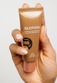 L'Oréal Paris GLOTION Natural Glow Enhancer in a squeeze tube; brown with a glossy finish, featuring white and black text, and a soft peach cap.