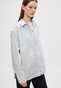 Ανοιχτό μπλε πουκάμισο με μακριά μανίκια και κάθετες ροζ ρίγες, με κουμπιά μπροστά, τσέπη στο στήθος και χαλαρή εφαρμογή. Το ύφασμα φαίνεται λείο και ελαφρύ.