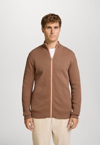 Brauner strukturierter Reißverschluss-Pullover mit orangefarbenem Reißverschlussdetail. Verfügt über einen Stehkragen und gerippte Bündchen, getragen über einem weißen Hemd.