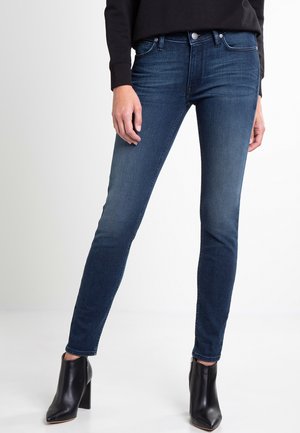 Žena nosící tmavě modré úzké džíny a černé kotníkové boty na podpatku stojí před bílým pozadím.