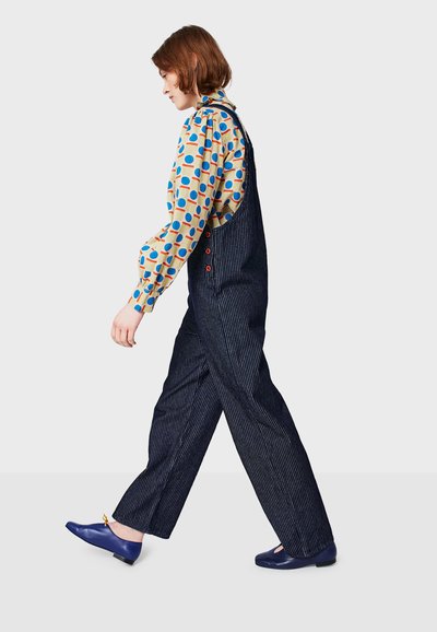 Blue and white striped overalls with side pockets, worn over a patterned long-sleeve shirt. Model wears blue shoes with an accent.