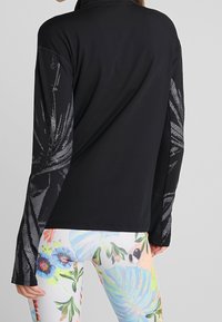 Kobieta w czarnej bluzce z długim rękawem z abstrakcyjnymi wzorami na rękawach i białych legginsach z kolorowymi tatuażami kwiatów i papug.