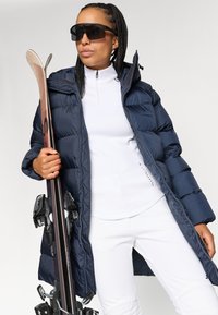 Geacă puffer bleumarin cu guler înalt, purtată cu un top alb cu fermoar și pantaloni albi. Ținând schiuri cu legături negre și purtând ochelari de soare mari și negri.
