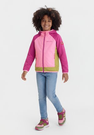 Barn med krøllet hår iført en jakke i pink, lilla og grøn, blå jeans og matchende sneakers, stående på ét ben med et smil.