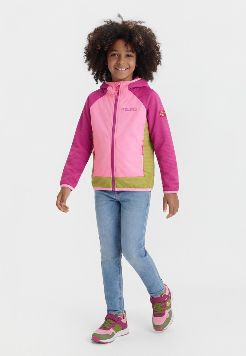 Παιδί με σγουρά μαλλιά που φοράει τζάκετ σε ροζ, μωβ και πράσινο χρώμα, μπλε τζιν και ταιριαστά αθλητικά παπούτσια, στέκεται στο ένα πόδι με ένα χαμόγελο.