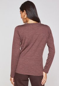 Camicia a maniche lunghe di colore bordeaux realizzata in un materiale morbido e texturizzato. Presenta un colletto rotondo e una forma aderente, abbinata a pantaloni marroni.