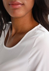 Haut blanc à manches courtes avec une texture lisse et un décolleté arrondi, présentant des détails de couture nets. Gros plan du vêtement sur un mannequin.