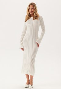 Dlouhý rukáv, pletené šaty s ribbed vzorem v off-white barvě, s přiléhavým střihem, bell rukávy a výstřihem do kulata. Textura je hladká a pružná.