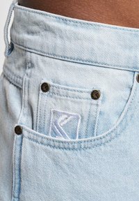 Pantalones cortos de mezclilla azul claro con un bolsillo frontal con herrajes de bronce y un logo blanco bordado en el bolsillo. Tela suave y texturizada.