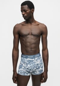 Homme musclé à la peau foncée portant un boxer Hollister bleu et blanc à motifs, debout devant un fond blanc uni, regardant vers la gauche.