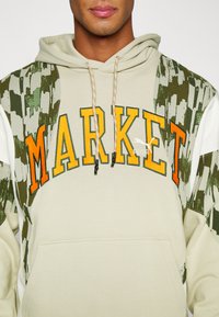 Felpa crema con la scritta "MARKET" in lettere arancioni e gialle. Presenta dettagli verdi con motivo mimetico e un cappuccio regolabile. Tasca frontale inclusa.