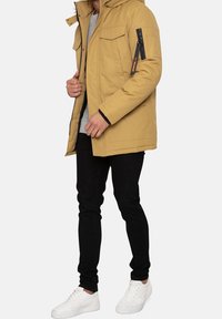Beige parka-jas met capuchon, voorzien van opgestikte zakken en een ritssluiting op de armzak, gedragen over een grijs shirt en zwarte broek, met witte sneakers.