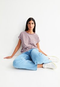 Frau mit langen dunklen Haaren sitzt auf dem Boden und trägt ein lila-weiß gestreiftes Shirt, hellblaue Jeans und weiße Sneaker vor einem weißen Hintergrund.