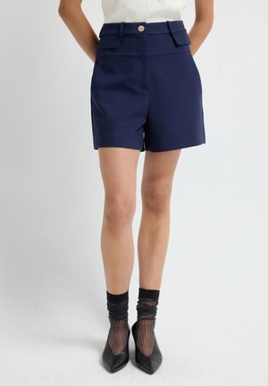 Femme portant un short taille haute bleu marine avec des poches à rabat, des chaussettes noires transparentes, des escarpins pointus noirs et un haut en tricot texturé blanc.