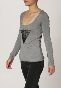 Haut gris à manches longues avec un col rond et un graphique triangulaire noir portant l'inscription "DEVINE QUI ?" sur le devant, fabriqué en tissu doux.